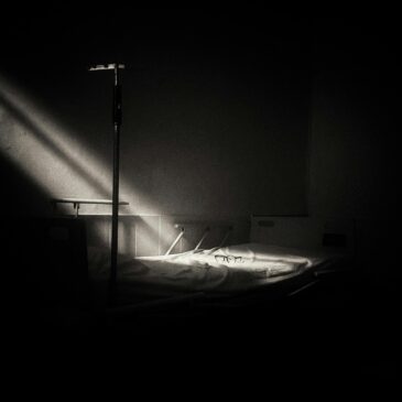 Black and white hospital room, dramatic light highlighting an empty bed, evokes solitude.